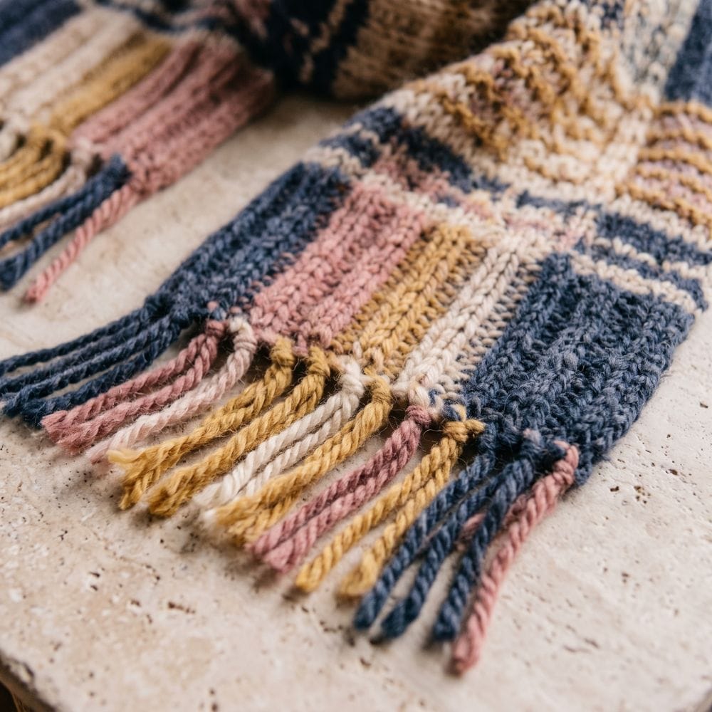 A close-up photo of a plaid knitted scarf with a fringe in navy, rose and mustard