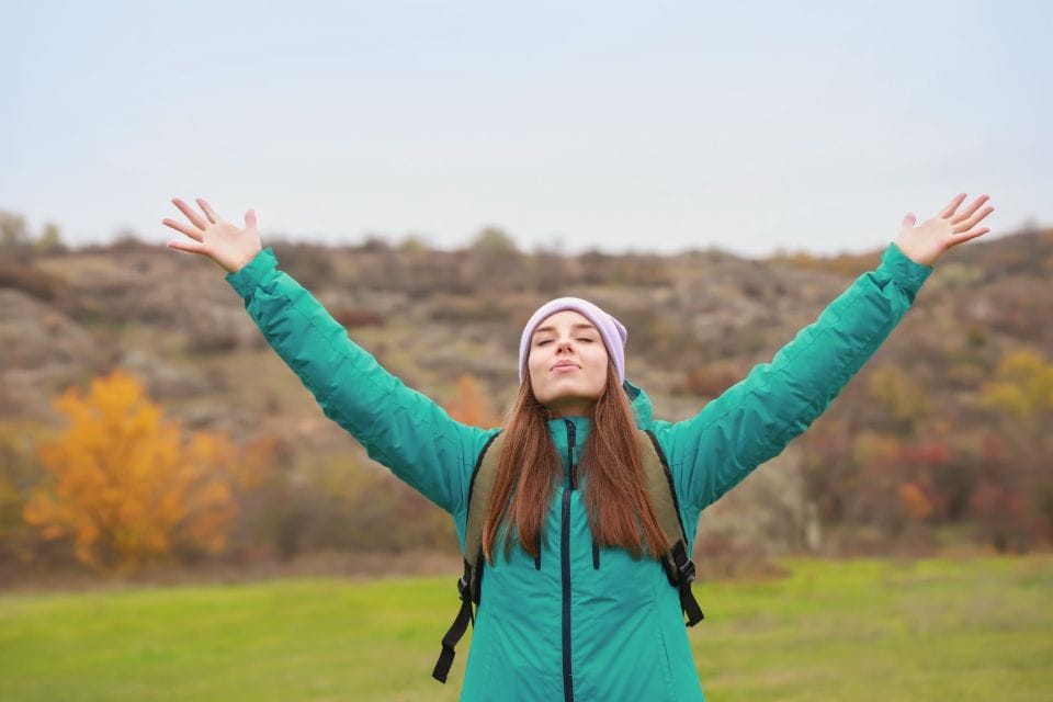 A hiker in a green jacket with both arms raised in a field