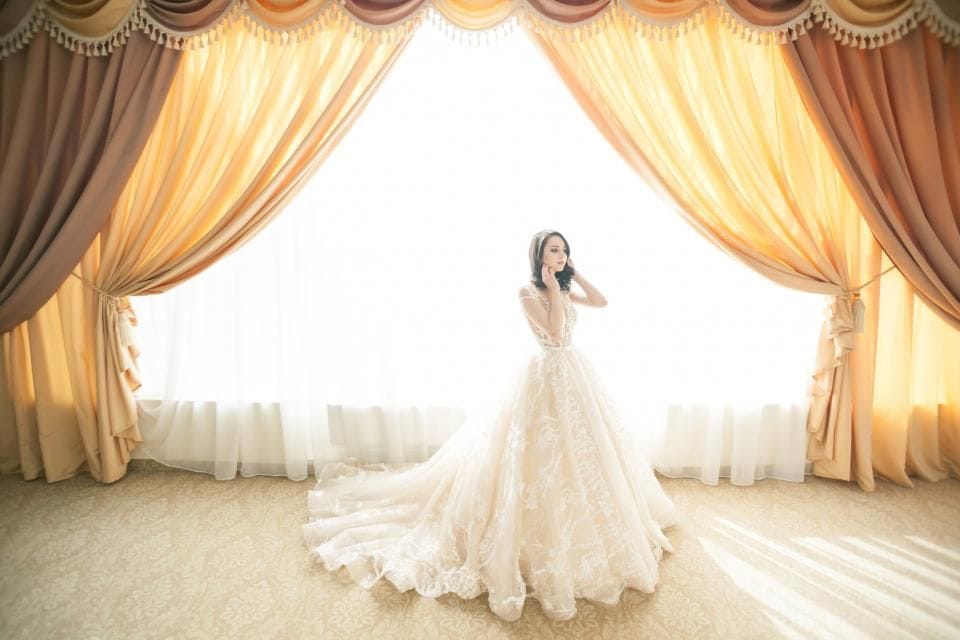 A bride in a full white gown standing between tall gold curtains in soft window light