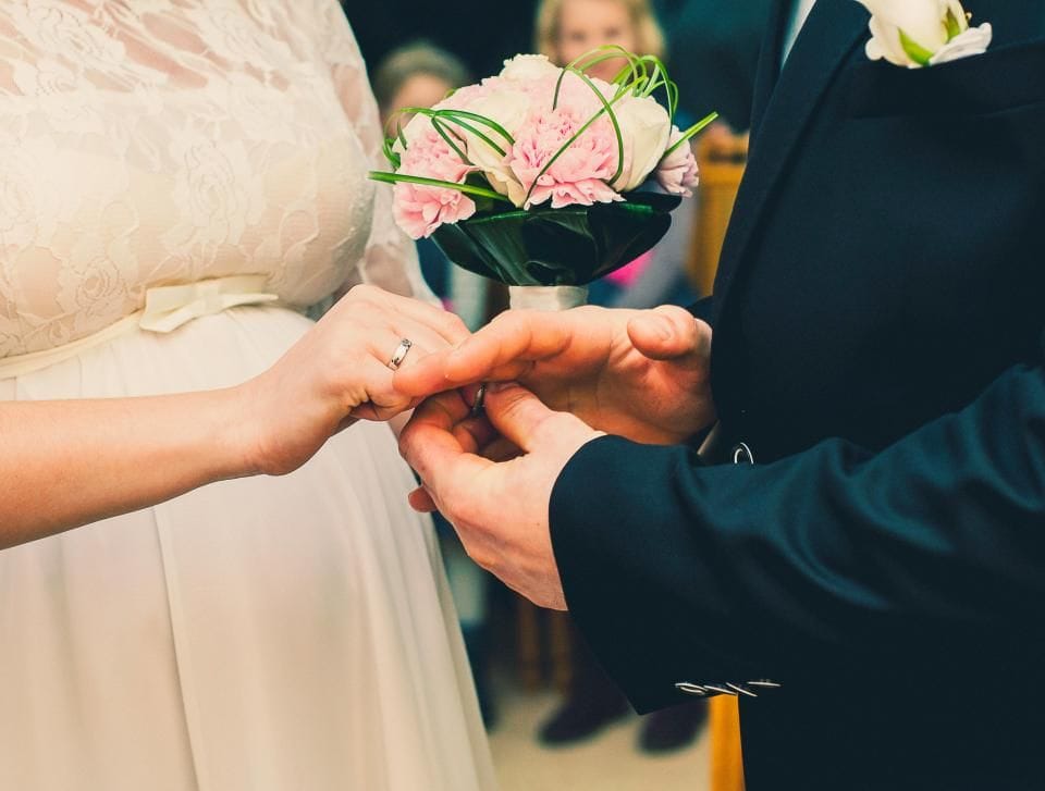 A groom sliding a ring onto the bride's finger, a pink bouquet between them