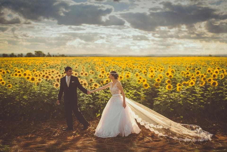 A bride and groom holding hands in a field of sunflowers under a cloudy sky, her long veil trailing behind