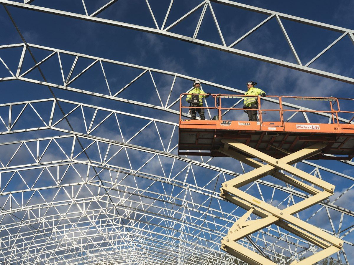 Two workers on a scissor lift under the steel roof trusses of a large single-storey building