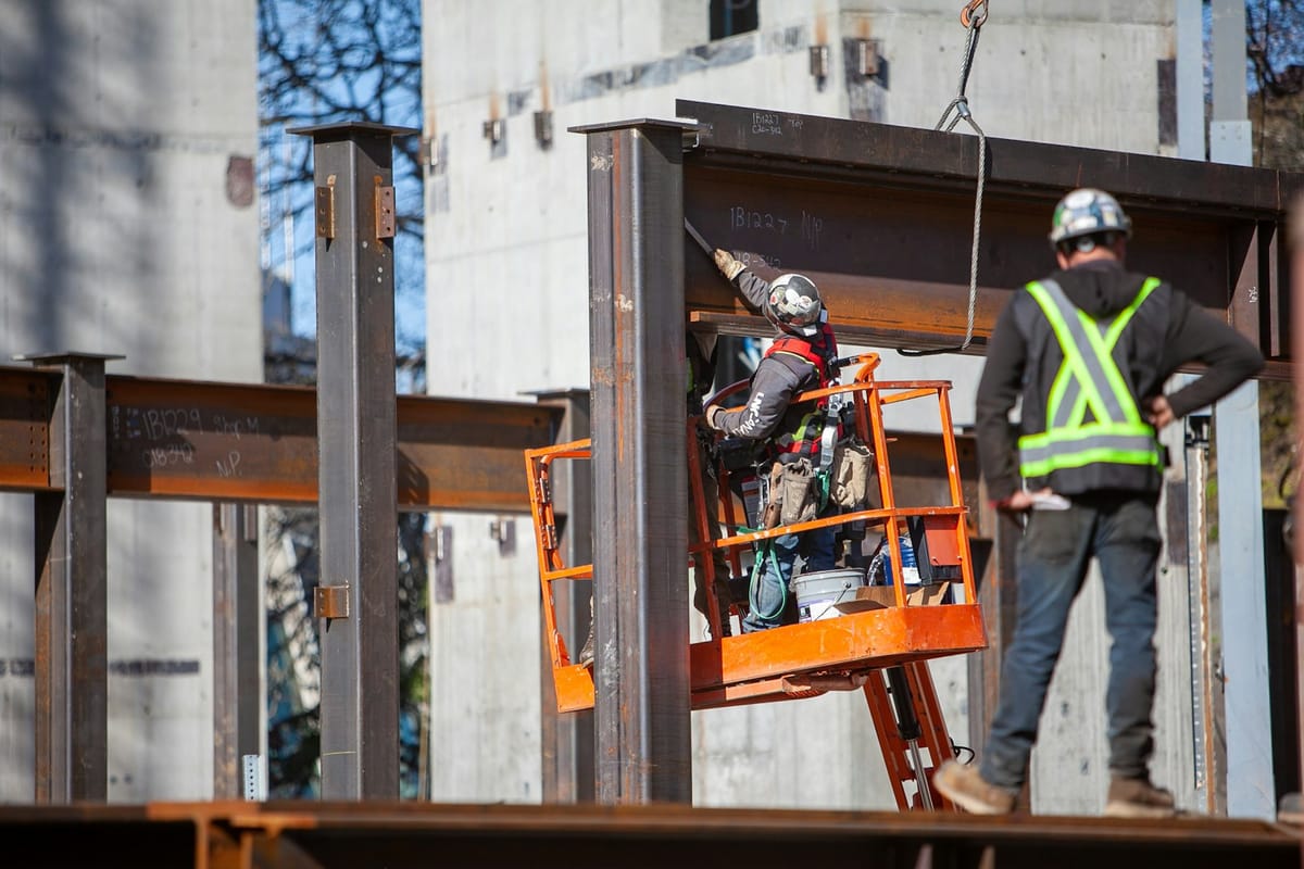 Ironworkers on a lift bolting up a steel frame, one in a high-visibility vest watching from the beam