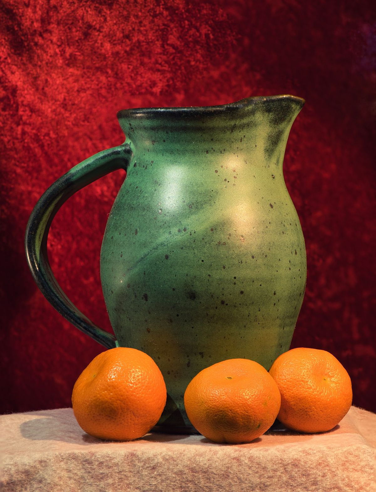 A green glazed pitcher with three oranges against a red backdrop