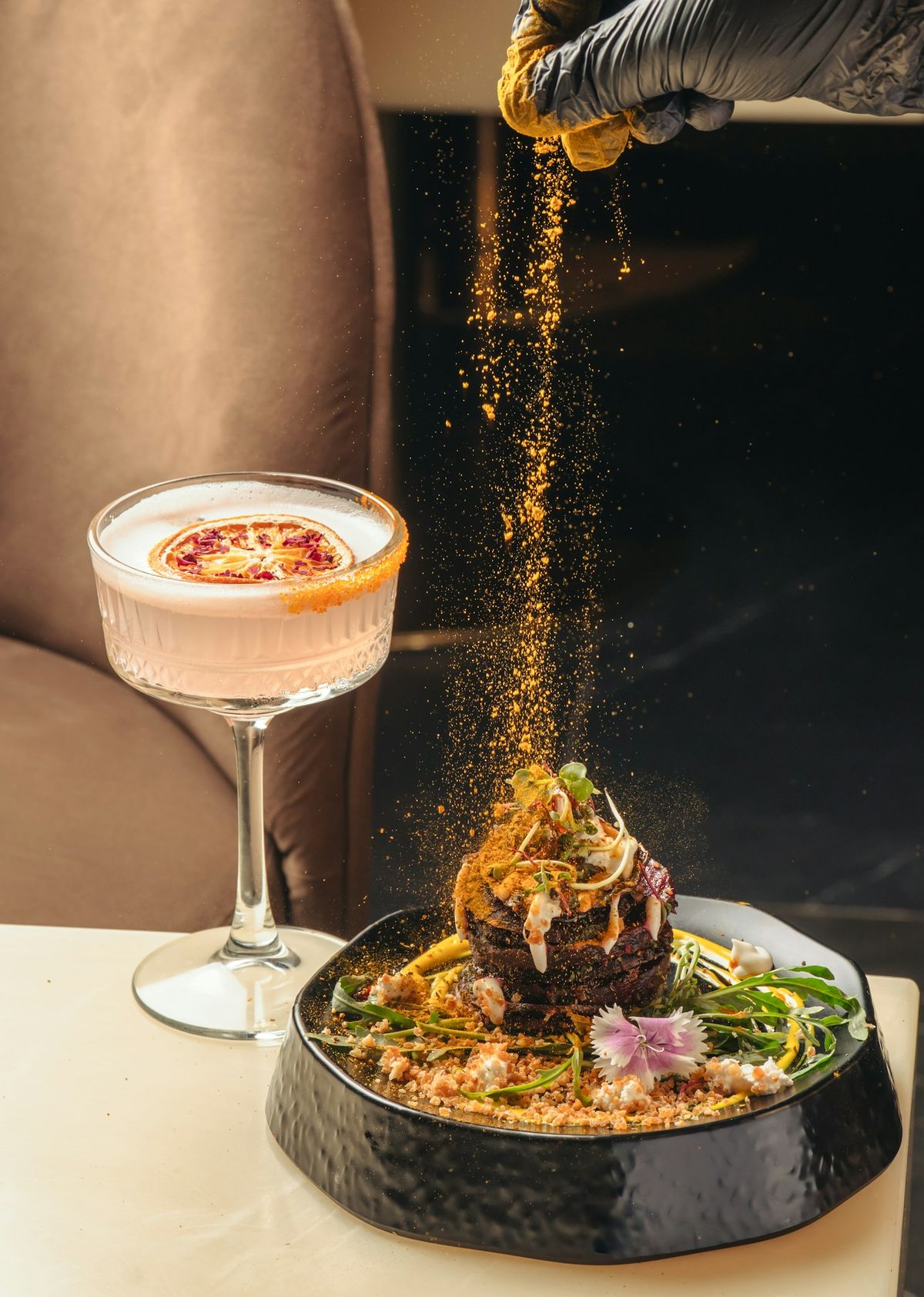 A chef sprinkling spice over a plated dish beside a cocktail