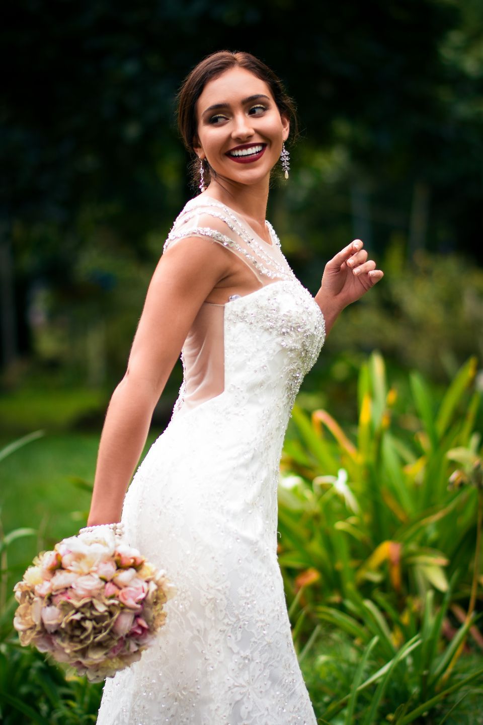 A bride in a lace gown laughing in a green garden, holding a bouquet